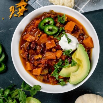 white bowl filled with pumpkin chili on a black countertop. the bowl is also filled with avocado slices, fresh cilantro, sour cream, and peppers. these ingredients are also scattered around the bowl.