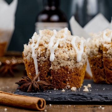 close up of an unwrapped gluten free pumpkin muffin with crumb topping and icing dripping down the sides. it rests on a black plate with whole spices beside it. more muffins are blurred in the background.