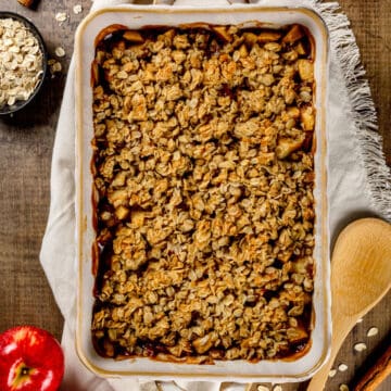 Closeup of a dairy free apple crisp in a ceramic baking dish on a wood tabletop. Oats, apples, a wood spoon, and cinnamon sticks surround the crisp.